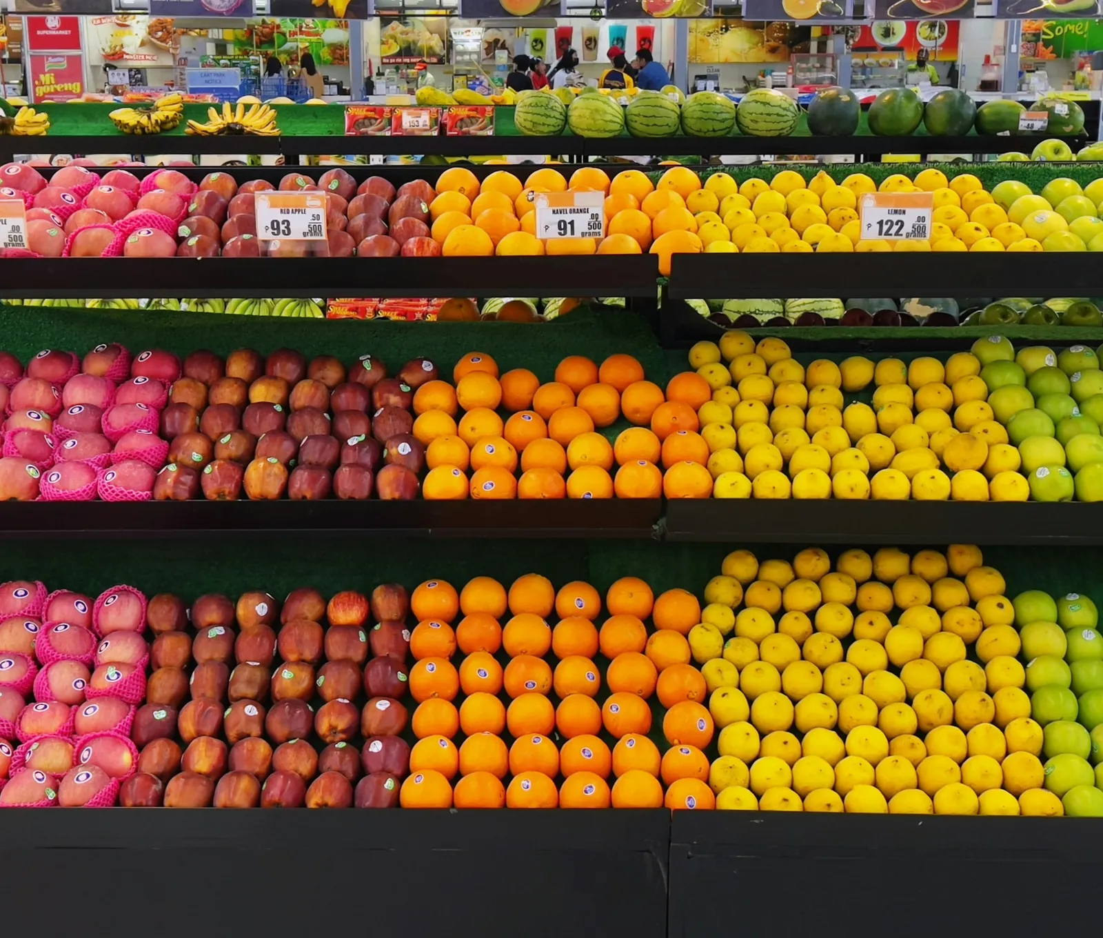 Rows of apples, oranges and lemons in a grocery store, with shoppers behind